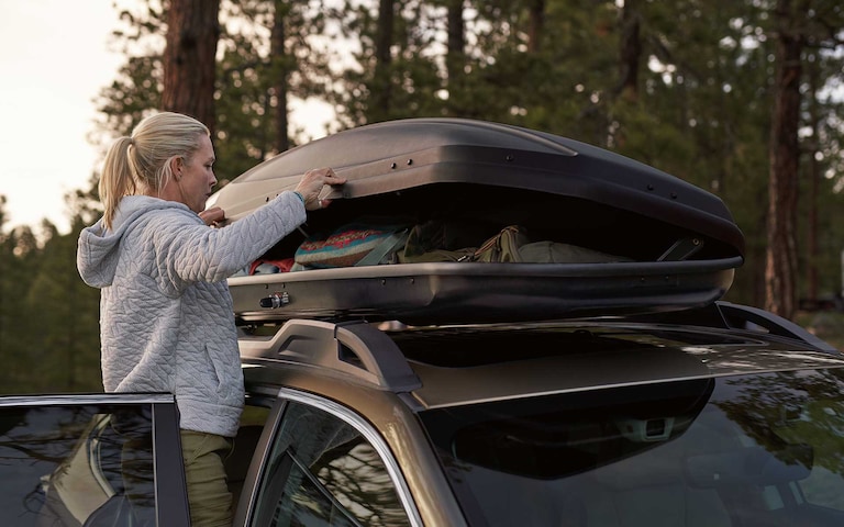 A woman opening a cargo container accessory mounted to the standard raised roof rails on a 2022 Subaru Outback.